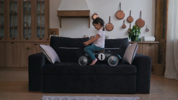 Little Girl Rides a Toy Car on the Sofa