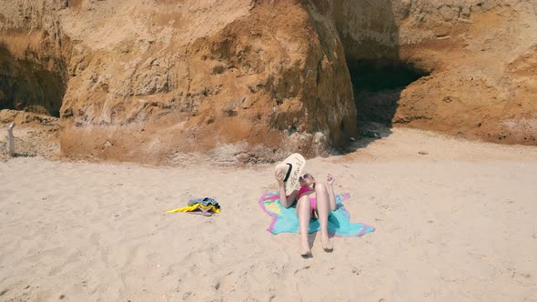 A Young Girl Lying on the Sand and Sunbathing Covering Her Face with a Big Hat alt