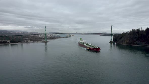 Red barge with pilot boat navigating under Lions Gate bridge with Vancouver city in background, Cana alt