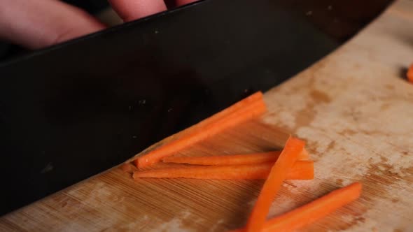 Black knife cuts carrot into thin slices in slow motion, on wooden chopping board. alt