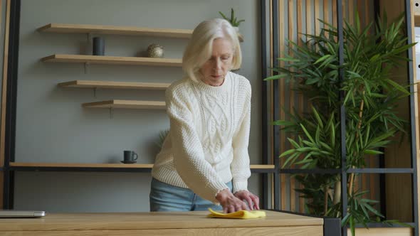 Elderly Woman Wipes Her Desk with a Rag. Concept of Homework and Household Management alt