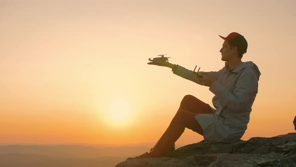 Man Sitting on Edge of Rock Cliff and Starts Drone From His Hand at Sunset alt