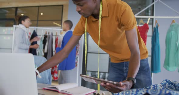 African american male designer standing using tablet and laptop in fashion studio alt