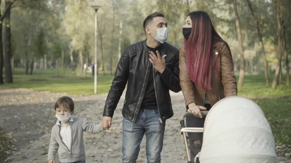 Loving Couple of Parents in Covid19 Face Masks Walking with Little Son and Infant in Baby Stroller alt