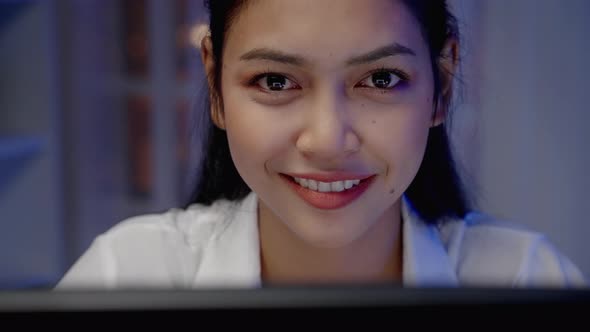 Close-up, Asian woman smiling in front of computer monitor at night.