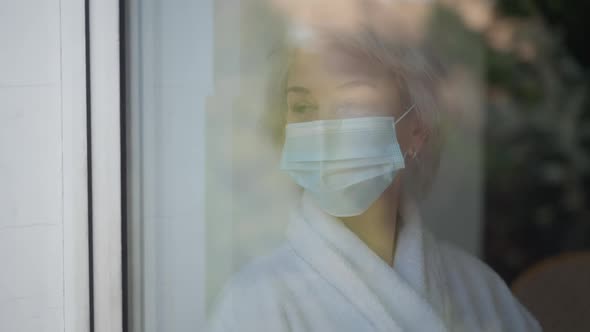 Shooting Through Window Sad Mature Woman in Covid19 Face Mask Standing Indoors Looking Out alt