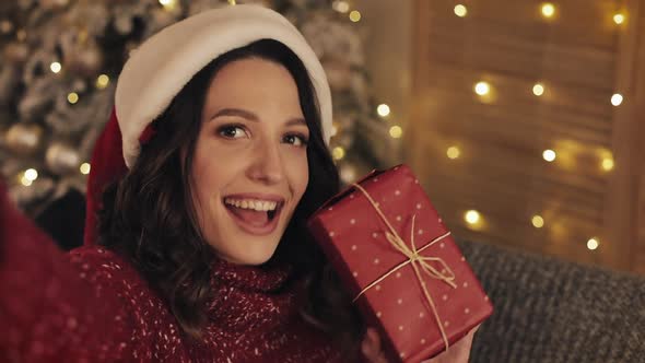 Young Smiling Pretty Brunette Woman Holding Present Box Sitting Near Christmas Tree Shining Lights alt