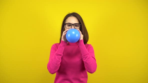 A Young Woman Inflates a Blue Balloon with Her Mouth on a Yellow Background. Girl in a Pink alt