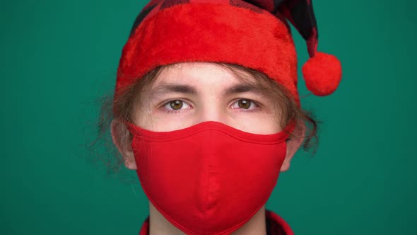 Portrait of Boy in Santa Hat Wears a Red Medical Mask Looking at Camera alt