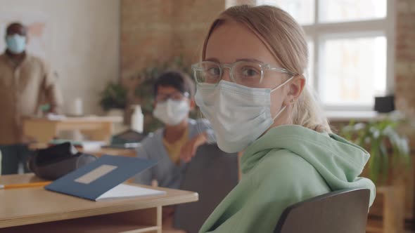 Blonde Schoolgirl in Disposable Face Mask Posing for Camera in Classroom alt