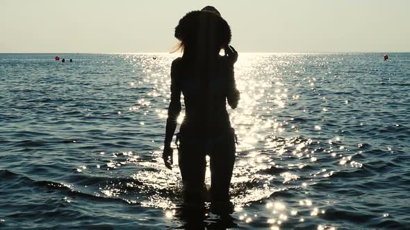 Silhouette Of Girl In Fashionable Hat Walking On The Sea Water On Sunset