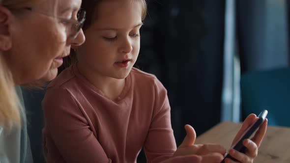 Side view of caucasian grandmother teaching granddaughter how to use  mobile phone in the kitchen. alt