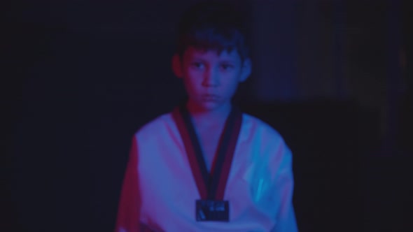 A Little Boy Doing Taekwondo in the Dark with Neon Lighting Walks to the Camera and Showing His alt