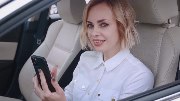 Beautiful Young Woman Sitting in a Car on Drivers Seat and Using Smartphone alt
