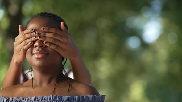 Closeup Young African American Woman Standing in Park As Friend Covering Eyes with Hands alt