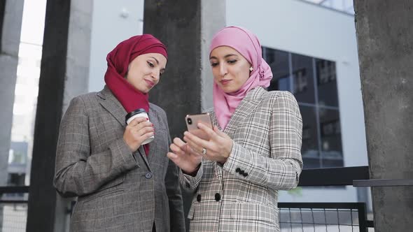 Two Young Muslim Women Wearing Hijab Headscarf Look at Phone, Walking Together Laughing Smile