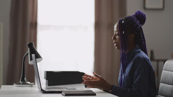 Black Woman is Consulting Online Sitting at Table with Laptop Looking at Web Camera and Answering alt