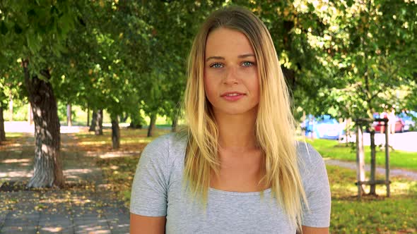 Young Pretty Blond Woman Looks To Camera with Serious Face - Park with Trees in Background alt
