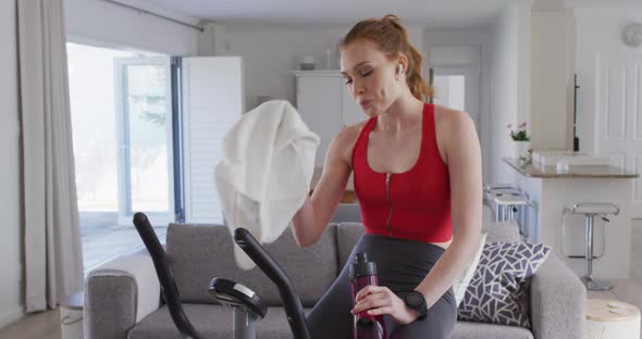 Woman drinking water and wiping her face with a towel while sitting on stationary bike at home alt