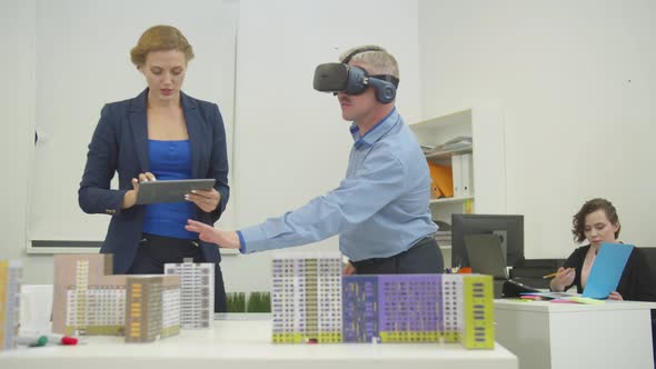 Woman Stands Next To Model of Houses with Tablet in Hands Talks with Colleague in Virtual Glasses in alt