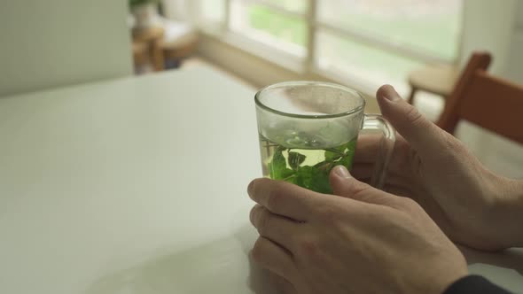 Man holding a glass of hot mint tea on a table in cozy apartment with garden alt