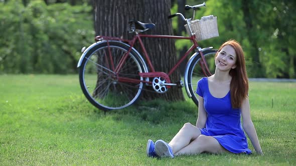 Redhead woman in blue dress sits on green grass with bike on background