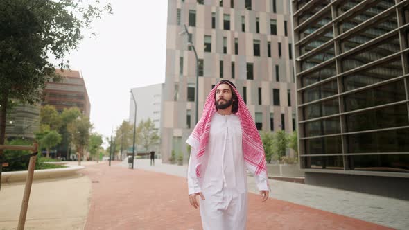 Successful Arab Man Sheikh in Traditional Clothes Looking at Skyscrapers While Walking City Street alt