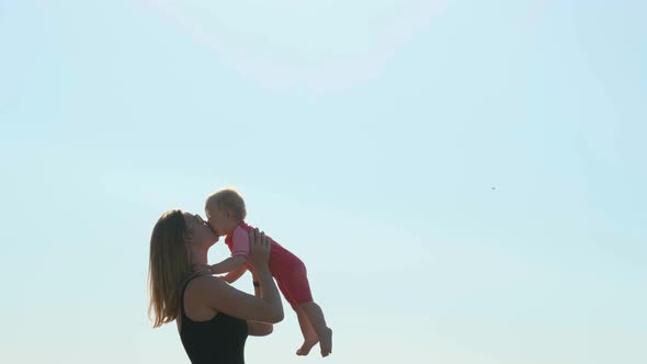 Sexy Mother in Black Closed Swimsuit on Seashore or Ocean Throws Her Daughter Up. Child in Pink alt