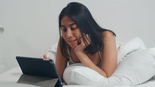 Young Asian woman using tablet checking social media feeling happy smiling while lying on bed.