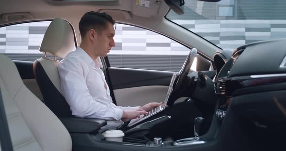 Portrait of Young Male Entrepreneur Sits in the Driver's Seat in a Car and Uses Laptop alt