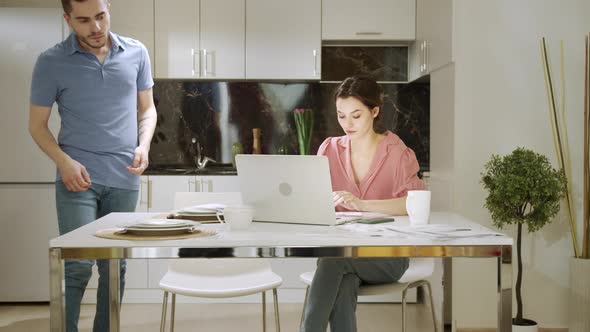 An Adult Woman is Sitting and Working at Home While Her Husband is Doing Household Chores alt