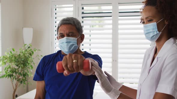 Mixed race female physiotherapist wearing mask helping senior exercise using dumbbells alt