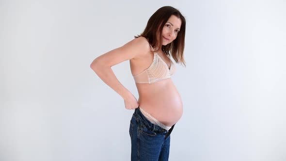 Pregnant woman in underwear puts on jeans in a white background