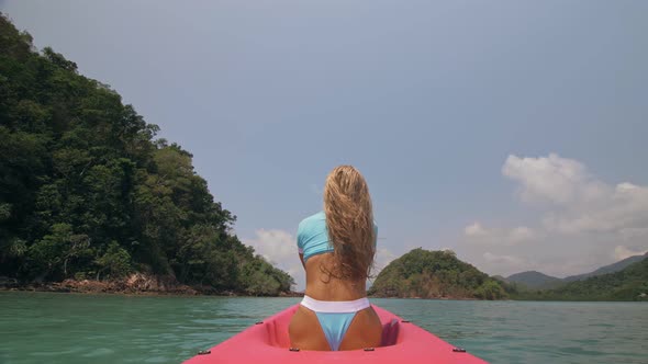 Sexy Young Woman in Blue Swimsuit Swings Feet in Water on Pink Canoe on Sea Against Hills Backside alt
