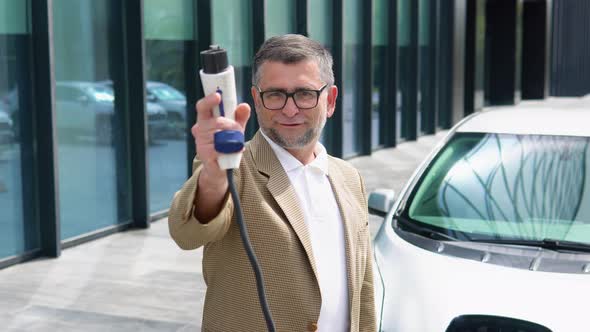 Older Man Stands with Charger Near His Electric Car alt