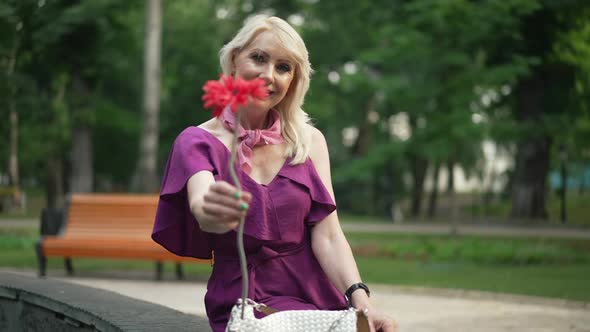 Medium Shot Positive Adult Woman Stretching Red Flower Looking at Camera Smiling Sitting at Fountain alt