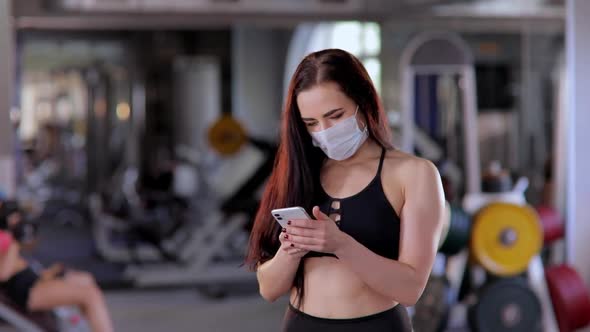 A Beautiful Athletic Woman Wearing a Protective Mask in the Gym Holds a Phone in Her Hand and Types