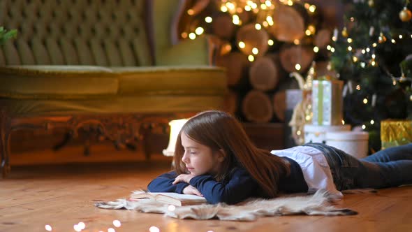 girl lies on the floor next to a book and dreams on the background lights and a festive tree