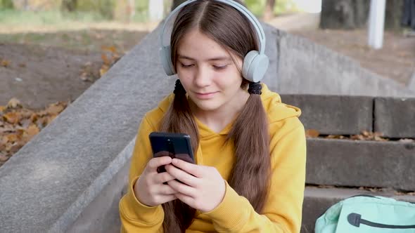 A Teenage Girl in Headphones Typing on a Smartphone and Smiling Texting with a Friend alt