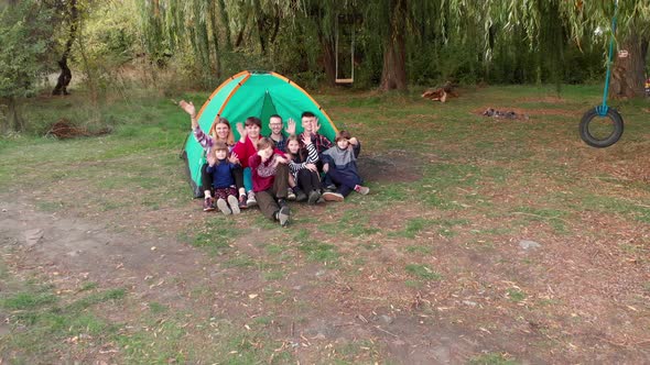 Pull Away Aerial Shot of Big Friendly Family Sitting on Grass and Wave Hands to Say Good Buy alt