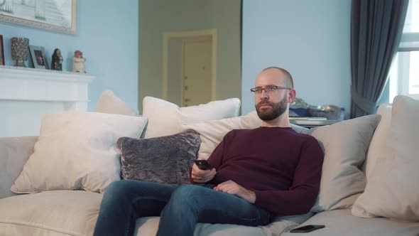 A young bearded man with glasses watching TV, sitting at home in a ...