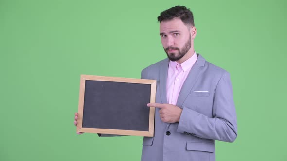 Happy Young Bearded Businessman Holding Blackboard and Giving Thumbs Up alt