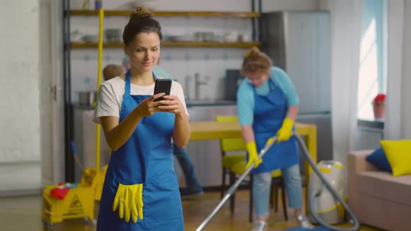 Positive Woman Janitor Using Mobile Phone During Break with Colleagues Cleaning Apartment on alt