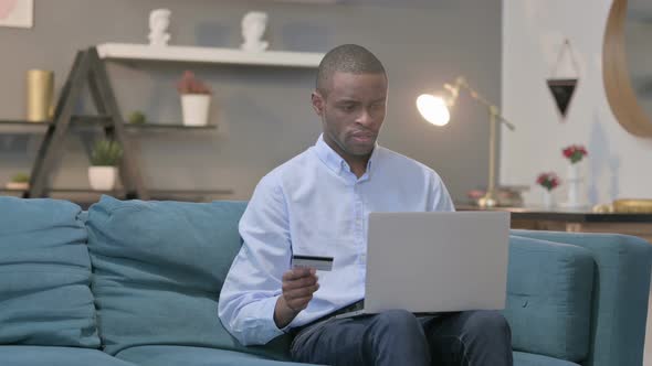 African Man Making Online Payment Failure on Laptop on Sofa alt