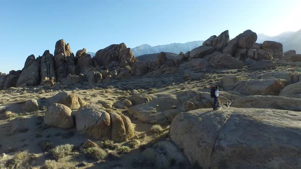 Aerial shot of a young man backpacker standing on a boulder with his dog in a desert mountain range. alt