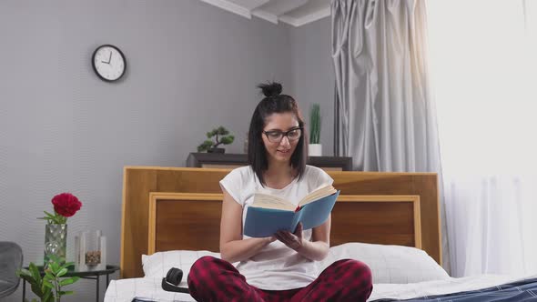 brunette in glasses which reading book ,sitting on comfortable bedroom bed near big window alt