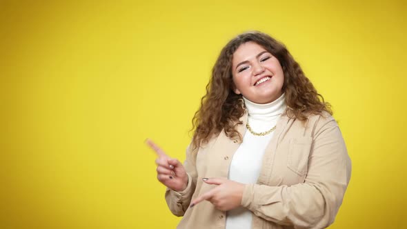 Joyful Smiling Plussize Woman Looking at Camera Pointing Aside Standing at Yellow Background alt