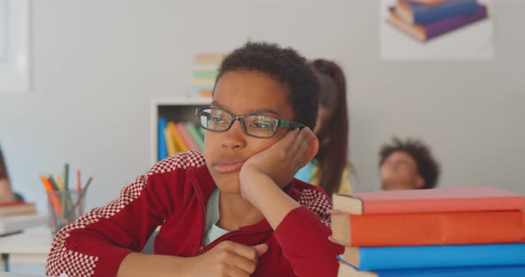 Tired African Schoolboy with Hand on Face Sitting at Desk in Classroom alt