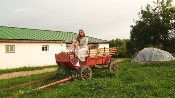 A Woman Sits in a Dress on an Old Rural Wagon Looking at the Camera