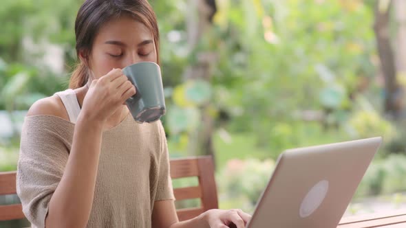 Freelance Asian woman working on laptop and drinking coffee sitting on table in the garden. alt